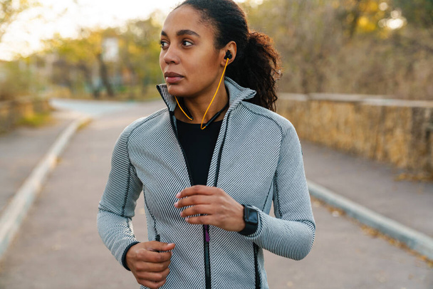 Beautiful african american sportswoman using earphones while running in park - Photo, image