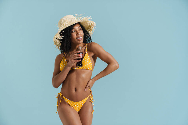 Black brunette woman in straw hat smiling while drinking soda isolated over blue background - 写真・画像