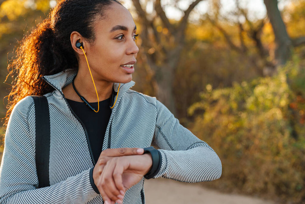 Pleased african american sportswoman in earphones using smartwatch in park - 写真・画像