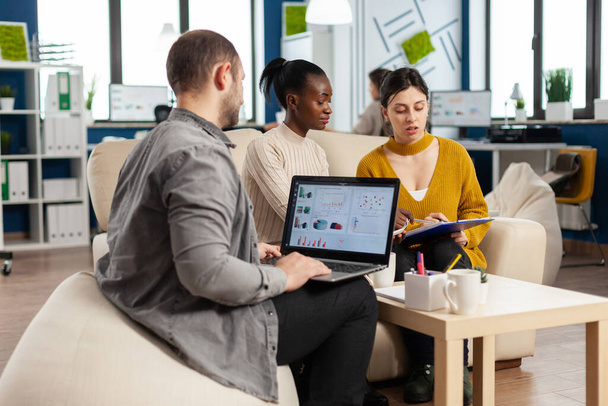 Empresario sosteniendo portátil con ghraphics financieros mientras diversos empleados hablan - Foto, Imagen