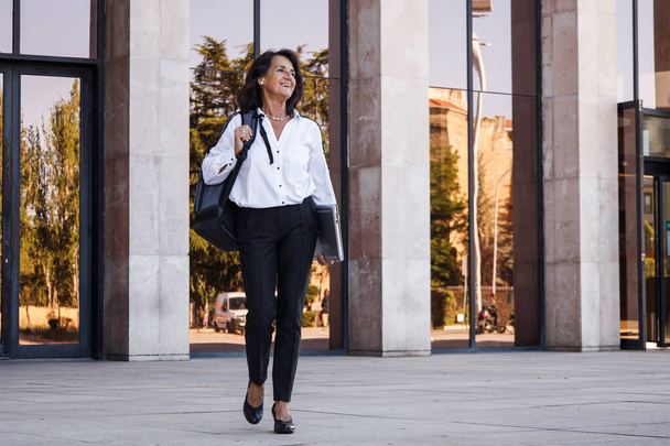 Hispánská zralá podnikatelka s batohem a bílou košilí telefonicky a se smíchem - Fotografie, Obrázek