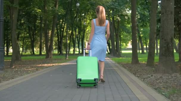 Young woman walking on park sidewalk with green suitcase on summer day. Travelling and vacation concept. - Footage, Video