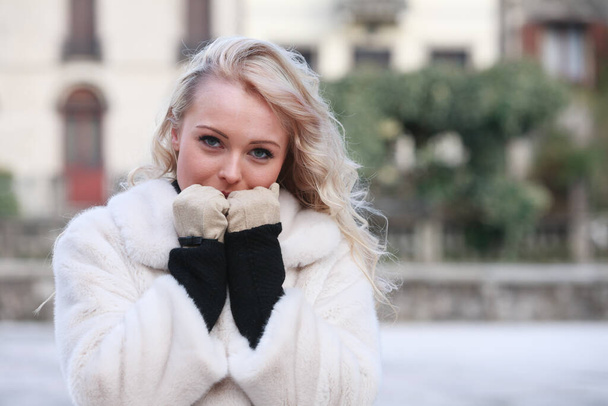 Young blond woman in stylish warm coat standing outdoors on a cold winter day holding her gloved hands to her face in an effort to keep warm as she looks at the camera - Photo, image