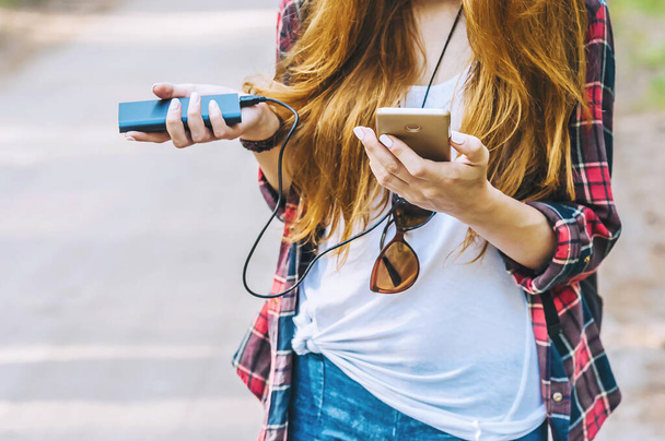Power bank and smartphone in the hands of a girl with red hair in a shirt in a cage with a black backpack, on the background of a forest road - Foto, imagen