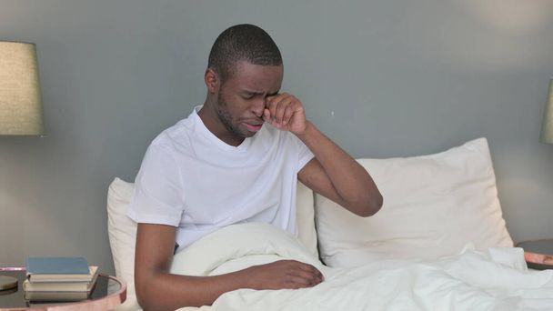 Расстроенный молодой африканский мужчина сидит в постели и плачет - Фото, изображение