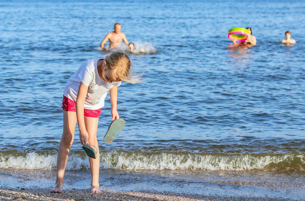 Kleines Mädchen von sechs Jahren, blond, steht am Strand, beugt sich vor und hält Hausschuhe in der Hand, Ganzkörperporträt eines Vorschulkindes im Meer - Foto, Bild