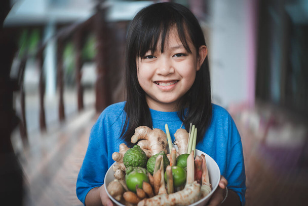 Ασιατικό κοριτσάκι κρατώντας ένα μπολ με βότανα περιλαμβάνουν πολλούς τύπους όπως τζίντζερ, galangal, λεμονόχορτο, kaffir lime, λεμόνι. Έννοια των βοτάνων για την αντι-ιική και ανοσία στο σώμα - Φωτογραφία, εικόνα