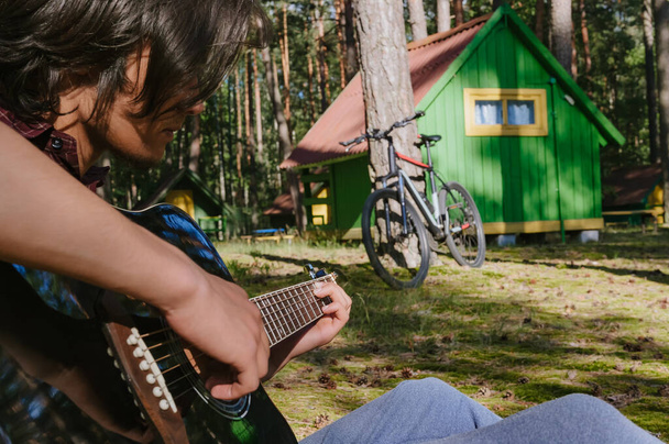 чоловік грає на гітарі. На тлі лісового будинку в природі
 - Фото, зображення