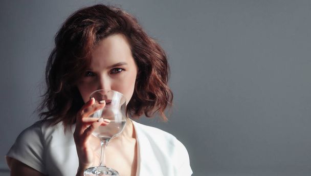 Close-up portrait of cute adult woman hold glass of water, drink and look at camera on dark background. Home photo of female with fresh aqua. Concept of water balance regulation and thirst quenching - Foto, afbeelding
