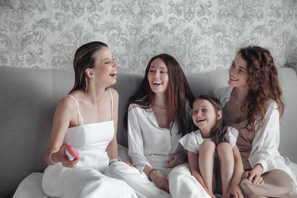 Three adult women and small girl in stylish casual white clothes spend time on bed, chat sweetly, tell stories and laugh. Friends met and spend their leisure time in hotel room. Concept of friendship - Фото, зображення