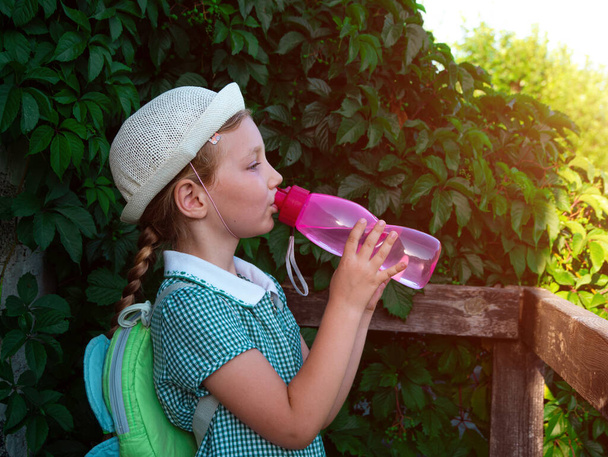 Linda niña de la escuela bebe agua de la botella rosa reutilizable al aire libre. Niño en sombrero disfruta de agua fría fresca en la calle verde de verano. Rehidratación corporal clima caliente, organismo humano activos líquidos mantenimiento - Foto, Imagen