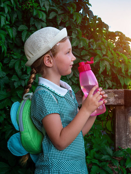 Симпатичная школьница пьет воду из многоразовой розовой бутылки на открытом воздухе. Ребенок в шляпе наслаждается пресной холодной водой на зеленой летней улице. регидратация организма жаркая погода, сохранение ликвидных активов человеческого организма - Фото, изображение