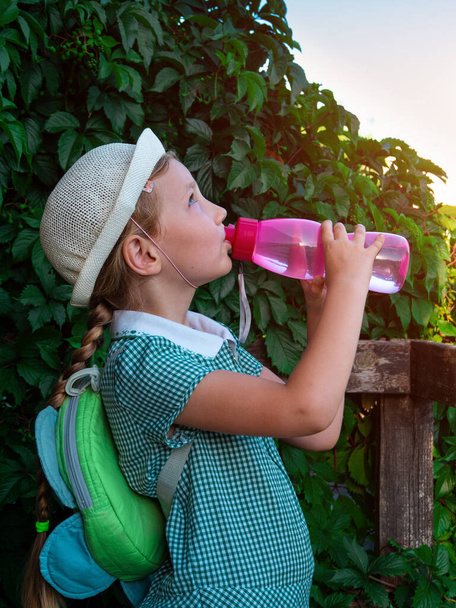 Cute school little girl drinks water from reusable pink bottle outdoor. Child in hat enjoys fresh cold water on green summer street. Body rehydration hot weather, human organism liquid assets keeping - Photo, Image