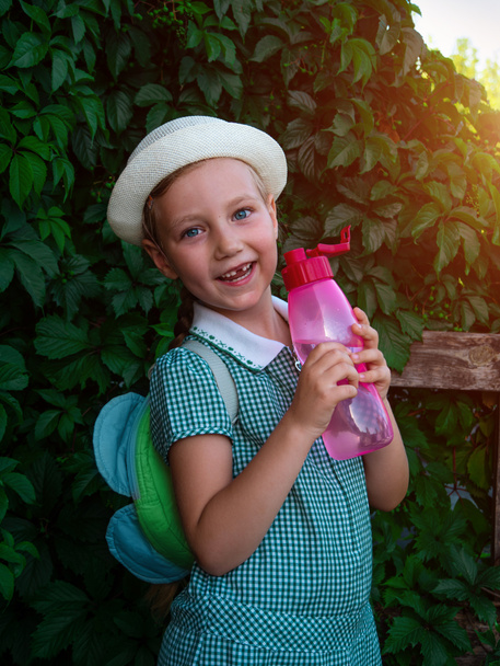 Cute school little girl drinks water from reusable pink bottle outdoor. Child in hat enjoys fresh cold water on green summer street. Body rehydration hot weather, human organism liquid assets keeping - Photo, Image