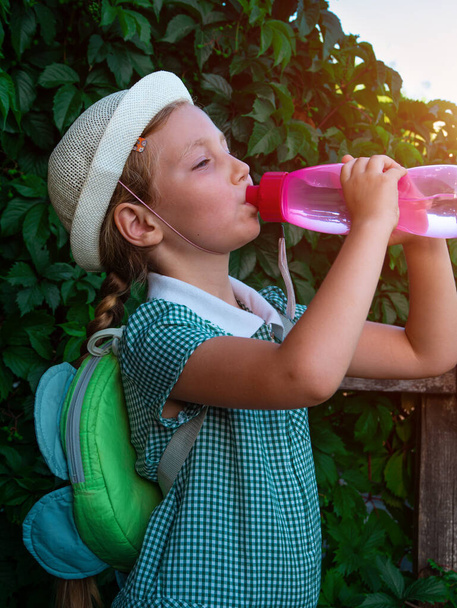 Cute szkoły dziewczynka pije wodę z wielokrotnego użytku różowej butelki na świeżym powietrzu. Dziecko w kapeluszu cieszy się świeżą zimną wodą na zielonej letniej ulicy. Nawadnianie ciała gorąca pogoda, organizm ludzki aktywa płynne utrzymanie - Zdjęcie, obraz