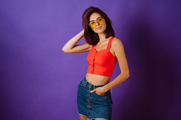 Beautiful young woman in a red top, skirt and yellow glasses posing on a purple background. Beauty and positive concept. - Фото, зображення