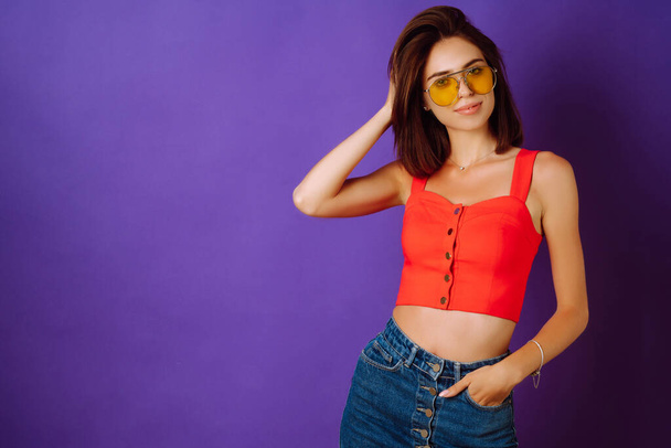 Beautiful young woman in a red top, skirt and yellow glasses posing on a purple background. Beauty and positive concept. - Фото, изображение