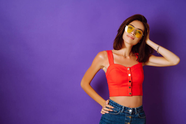 Beautiful young woman in a red top, skirt and yellow glasses posing on a purple background. Beauty and positive concept. - Fotoğraf, Görsel