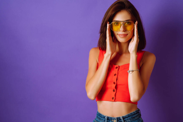 Beautiful young woman in a red top, skirt and yellow glasses posing on a purple background. Beauty and positive concept. - Fotografie, Obrázek