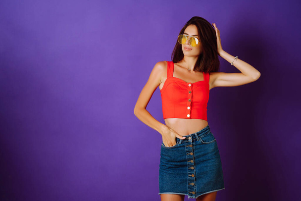 Beautiful young woman in a red top, skirt and yellow glasses posing on a purple background. Beauty and positive concept. - Valokuva, kuva