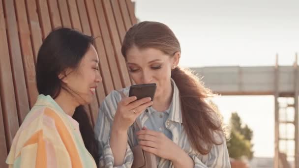 Pas-up záběr dvou radostných přítelkyň nahrávat vtipné hlasové vzkazy na smartphone a smát se stojící venku v letní den - Záběry, video