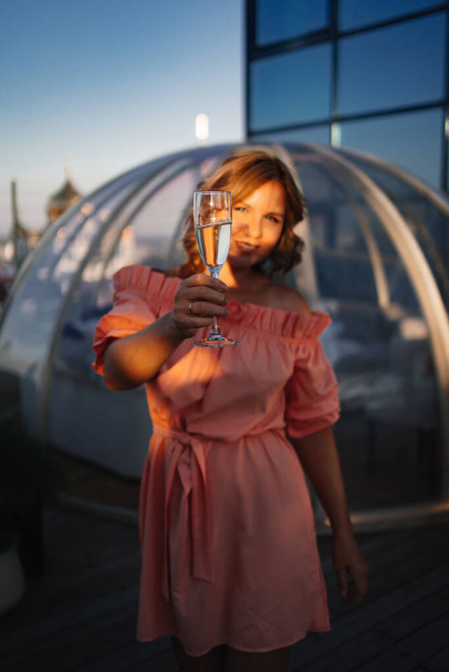 at sunset. beautiful woman in a pink dress on the roof with a glass of champagne - Фото, изображение