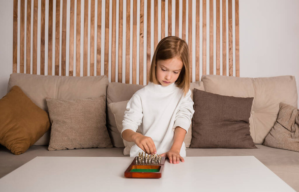 una niña rubia está sentada en una mesa y jugando al ajedrez en una habitación - Foto, imagen