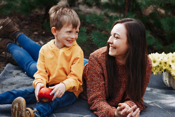 Family picnic: young beautiful caucasian woman with her little child lying on the blanket, rest, and having fun in the park. Happy mother with a son in sweaters in the pine forest. Motherhood concept. - Photo, Image
