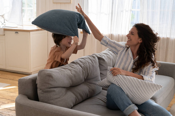 Fröhliche Mutter mit Vorschulkind kämpft gegen Kissen im Wohnzimmer. Lustige Familienaktivitäten zu Hause auf - Foto, Bild