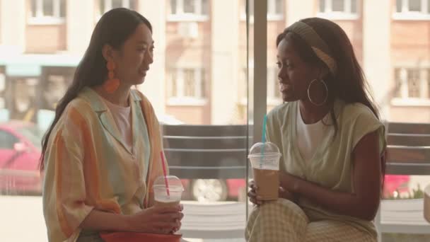 Foto en la cintura de dos jóvenes mujeres multiétnicas sentadas junto a la ventana en la cafetería en el caluroso día de verano, bebiendo café helado de vasos de plástico y charlando - Metraje, vídeo
