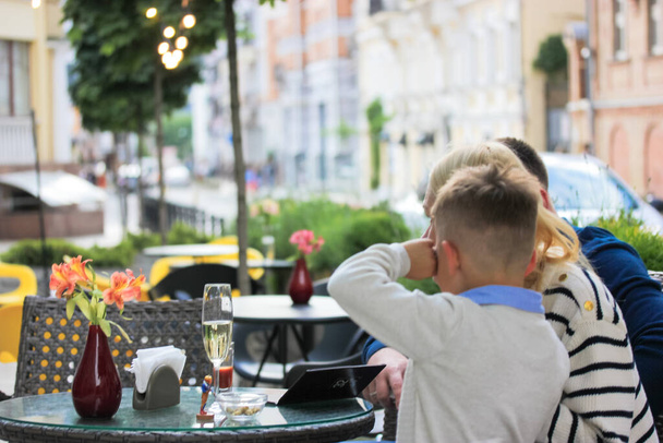 A family with a boy relaxing on cozy terrace on a warm summer day. Mother, father, son having breakfast in a morning. A glass of champagne on a table in a cafe, restaurant in a big city. Urban scene. - Photo, Image