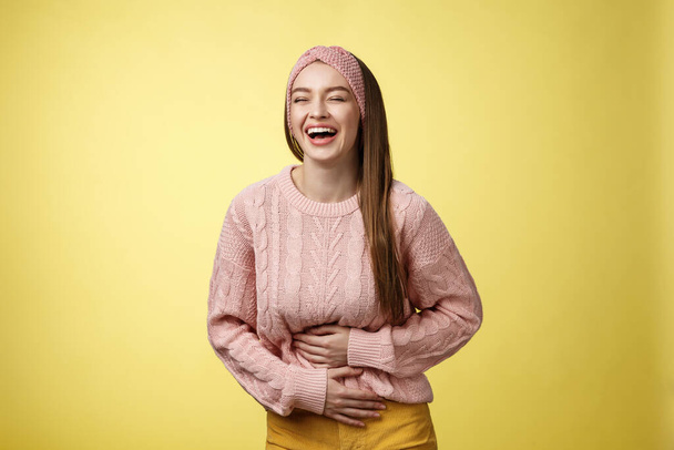 Задоволена і розважаюча молода європеїстка студентка сміється, поки живіт болить від посміхаючись пальцями на животі, насолоджуючись переглядом смішної комедії над жовтою стіною. - Фото, зображення