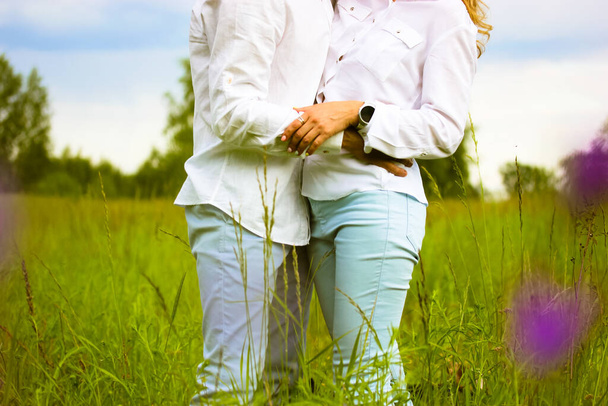 Hermosa pareja enamorada abrazando suavemente de pie en un prado verde en flor en verano. Esposo, esposa con camisas blancas mostrando sus sentimientos el uno por el otro. Devoción, pureza, armonía, concepto de amor. - Foto, imagen