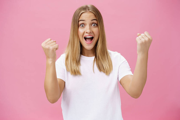 Excited cheerful and optimistic charming woman with fair hair in white t-shirt raising fists in victory and triumph yelling yes in success and amazement standing supportive against pink background - Photo, Image
