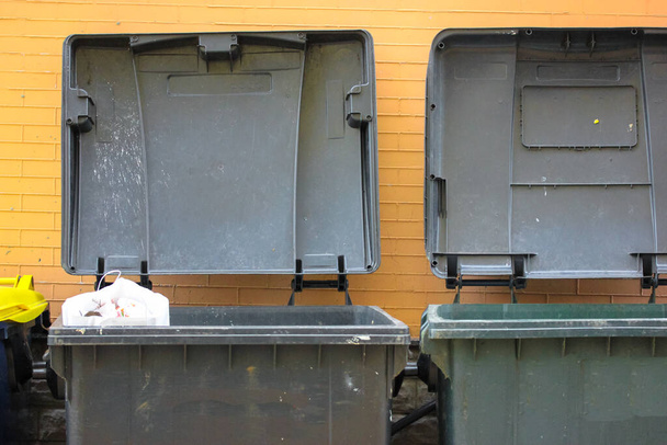 Пластиковые большие мусорные баки с крышкой и мусор внутри к кирпичной оранжевой стене. Большой зеленый или серый пластиковый мусорный контейнер на городской улице. Отходы мусорные баки. Ответственное отношение к окружающей среде. - Фото, изображение
