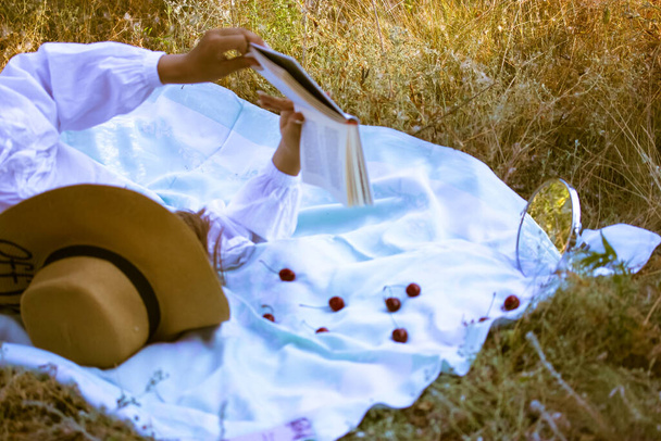 Una mujer con un sombrero de paja yace sobre un cuadros con un libro en sus manos. Leyendo literatura al aire libre sobre una hierba amarilla seca en un parque. Vacaciones, recreación, afición, concepto de vacaciones de verano. Cerezas rojas maduras. - Foto, imagen