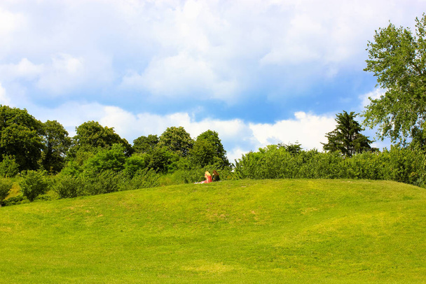 People are sitting relaxing on a lawn on a small hill in a countryside, public park, botanical garden. Green grass, trees against a blue cloudy sky before rain on a summer day. Amazing landscape.  - Photo, Image