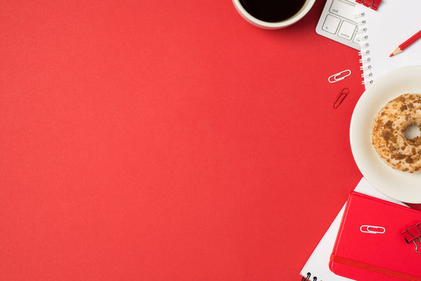 Vista superior de la foto del lugar de trabajo clips de teclado lápiz rojo copybooks rojo taza de café y plato con donut acristalado decorado sobre fondo rojo aislado con copyspace - Foto, imagen