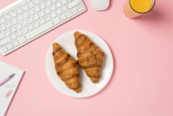 Overhead photo of keyboard computer mouse glass of orange juice notepad with pen paper clips and plate with two croissants isolated on the pink backdrop - Φωτογραφία, εικόνα