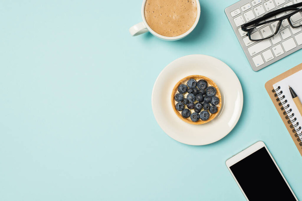 Vista superior de la foto de las gafas en la placa del bloc de notas del teclado con el teléfono inteligente del pastel de arándanos y la taza de café con leche espumosa en el fondo azul pastel aislado con copyspace - Foto, Imagen