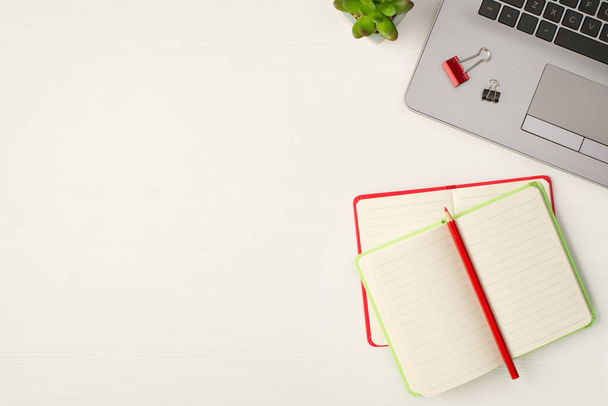 Foto von oben von Ordnerclips auf Laptop-Blumentopf-Bleistift auf roten und hellgrünen offenen Notizbüchern auf isoliertem weißem Holztischhintergrund mit Kopierraum - Foto, Bild