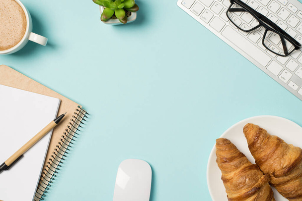 Horní pohled fotografie pracovních brýlí na klávesnici myš rostlina šálek pěnivé kávy pero plánovače a deska s croissanty na izolované pastelové modré pozadí s prázdným prostorem - Fotografie, Obrázek