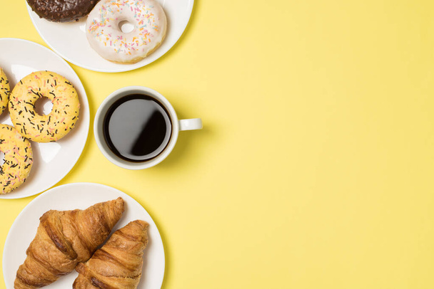 Top view photo of cup of coffee and three plates with different color donuts and croissants on isolated pastel yellow background with empty space - Photo, Image