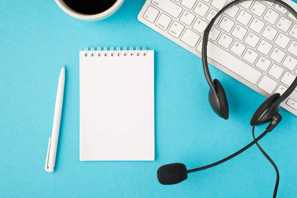 Фотографія чорного гарнітура з мікрофоном на білій клавіатурі чашки кави білої ручки та відкритого щоденника на ізольованому пастельно-блакитному фоні з порожнім простором
 - Фото, зображення