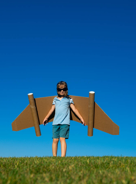 Child boy with paper wings against blue sky. Kid with toy jetpack having fun in spring green field outdoor. Freedom carefree and kids imagination dream concept. - Foto, Imagem