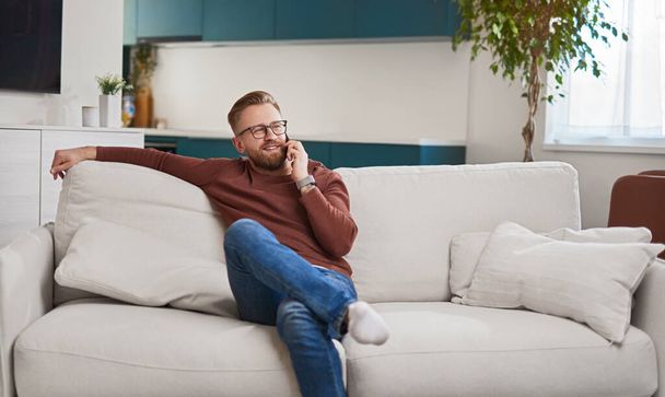 Relaxed man speaking on smartphone on sofa - 写真・画像