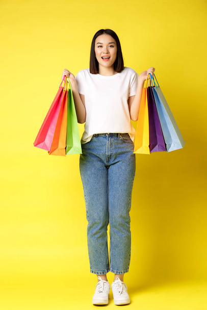 Plná velikost portrét krásné asijské dívky jít nakupovat, drží papírové tašky z obchodů a usmívá se, stojí v džínách a bílé tričko přes žluté pozadí - Fotografie, Obrázek