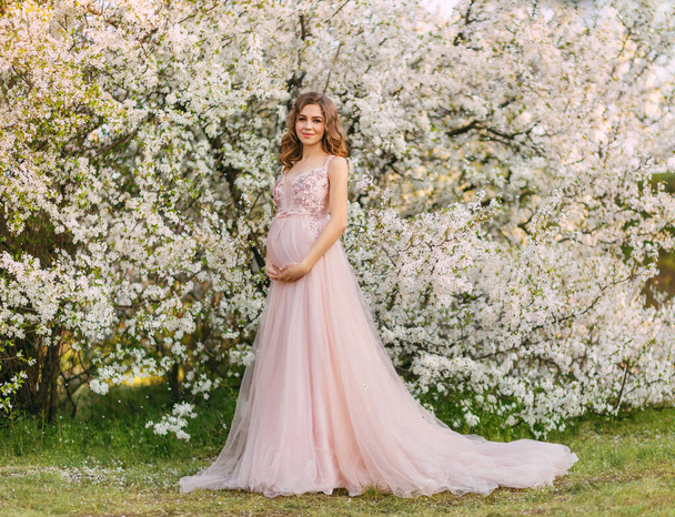 Young beautiful happy joyful pregnant woman in light pink long evening dress posing. background of blooming trees. Garden green leaves white flowers. Girl model. Elegance pregnancy spring fresh nature - Photo, Image