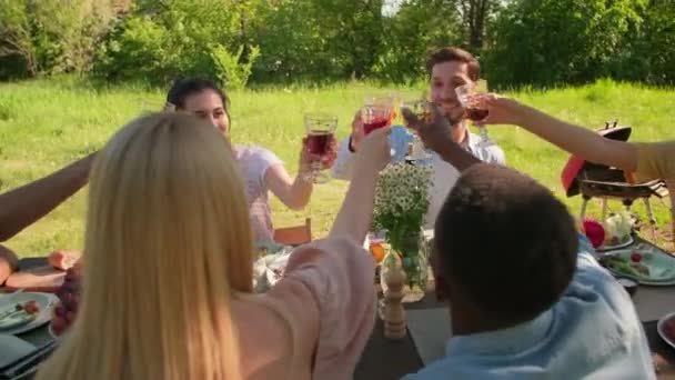 Tiro de mano de hombres y mujeres adultos jóvenes modernos que pasan tiempo juntos en el parque sentados en la mesa tintineo vasos con bebidas - Imágenes, Vídeo