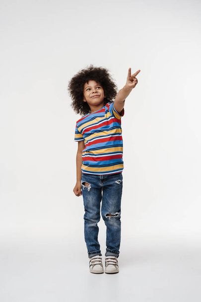 Happy preschooler african boy standing over white wall, having fun, full length, showing peace gesture - Photo, Image